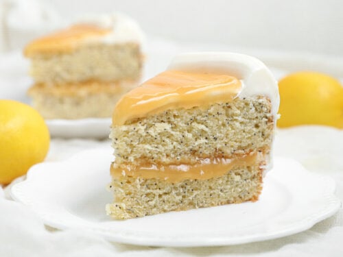 A slice of Lemon Poppy Seed Cake with creamy white frosting and yellow glaze sits on a white plate, surrounded by whole lemons and another tempting slice in the background.