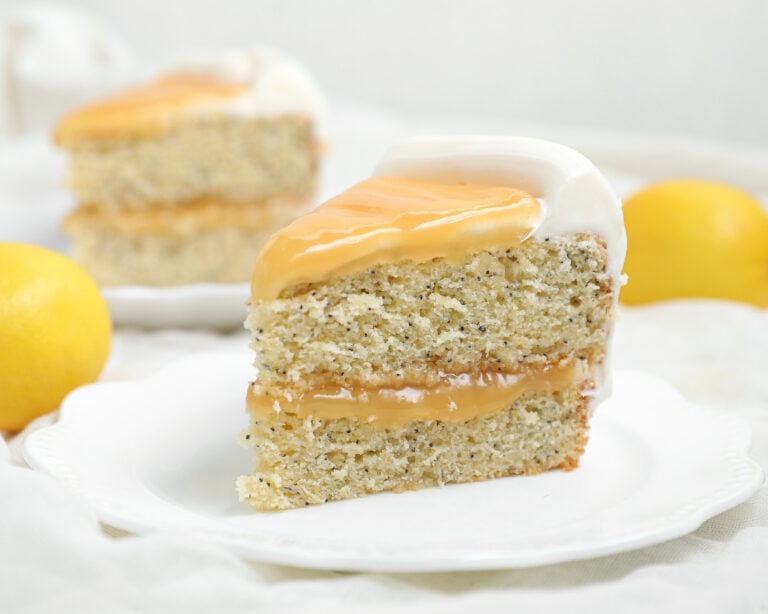 A slice of Lemon Poppy Seed Cake with creamy white frosting and yellow glaze sits on a white plate, surrounded by whole lemons and another tempting slice in the background.
