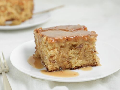 A slice of moist Pineapple Sticky Toffee Pudding Cake with luscious pineapple toffee sauce on top sits on a white plate, with a fork nearby. Another slice of cake is visible in the blurred background.