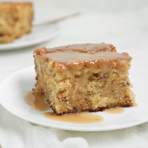 A slice of moist Pineapple Sticky Toffee Pudding Cake with luscious pineapple toffee sauce on top sits on a white plate, with a fork nearby. Another slice of cake is visible in the blurred background.