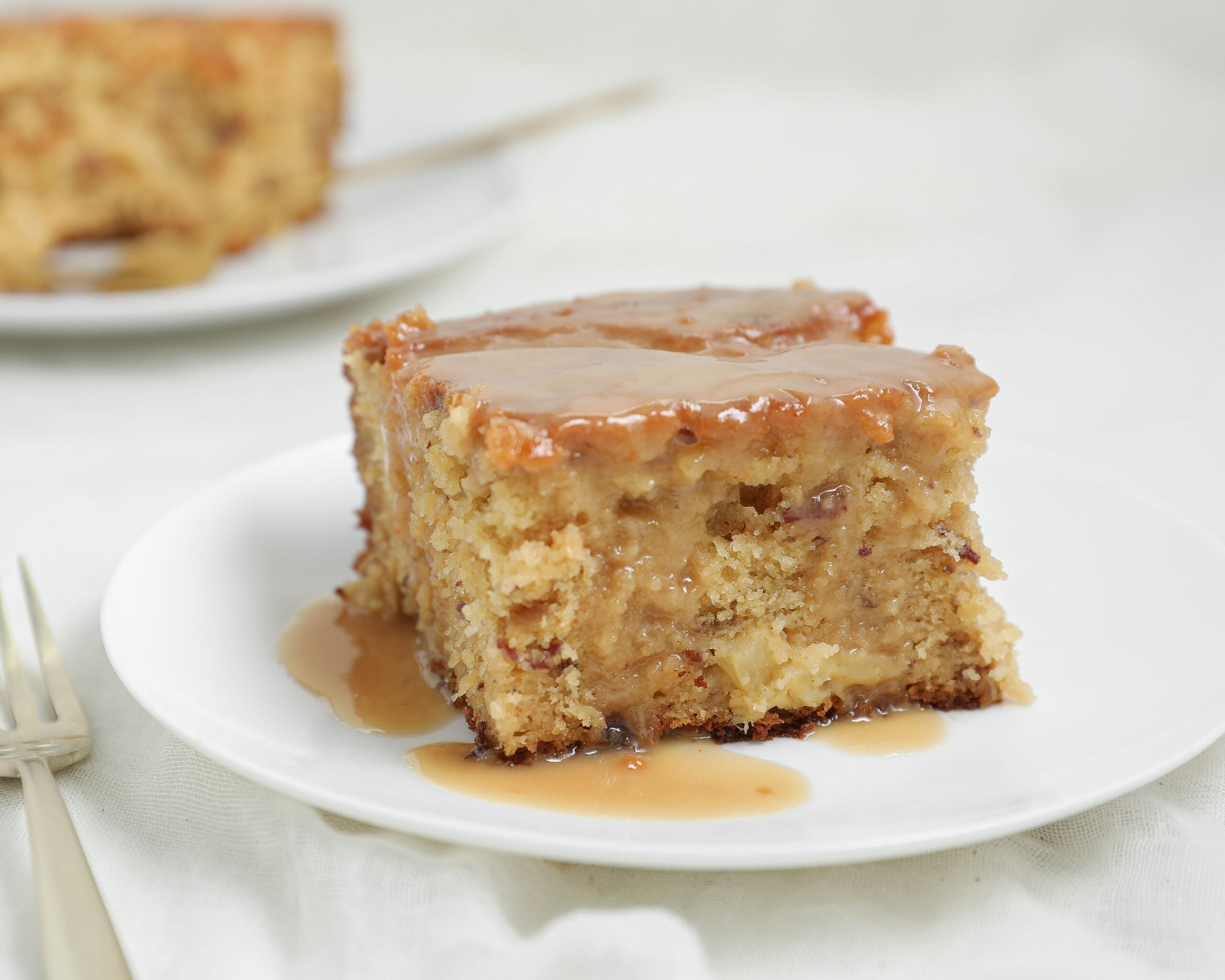 A slice of moist Pineapple Sticky Toffee Pudding Cake with luscious pineapple toffee sauce on top sits on a white plate, with a fork nearby. Another slice of cake is visible in the blurred background.