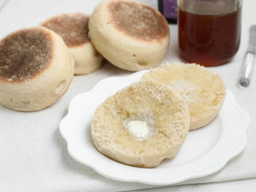 Split sourdough English muffin with melting butter on a white plate, with more English muffins, a jar of honey, and a butter knife in the background.