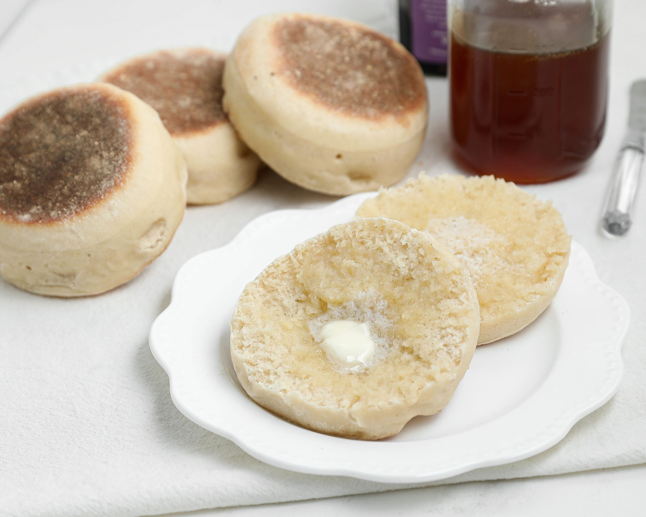 Split sourdough English muffin with melting butter on a white plate, with more English muffins, a jar of honey, and a butter knife in the background.