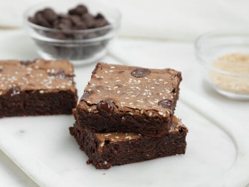 Two Dark Chocolate Tahini Brownies topped with sesame seeds, stacked on a white marble surface. In the background, small glass bowls hold chocolate chips and a light-colored ingredient.