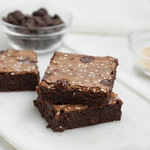 Two Dark Chocolate Tahini Brownies topped with sesame seeds, stacked on a white marble surface. In the background, small glass bowls hold chocolate chips and a light-colored ingredient.