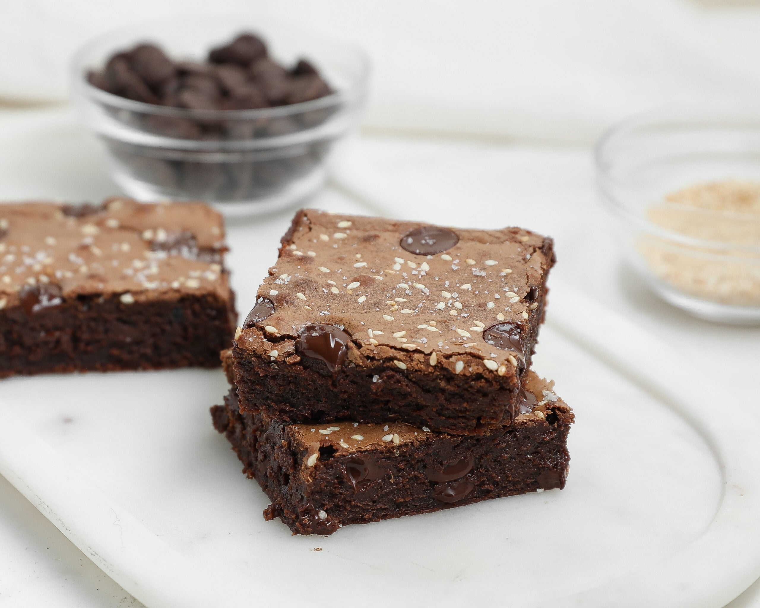 Two Dark Chocolate Tahini Brownies topped with sesame seeds, stacked on a white marble surface. In the background, small glass bowls hold chocolate chips and a light-colored ingredient.