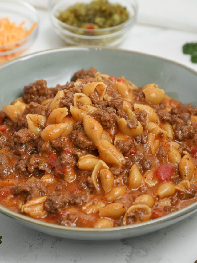 Bowl of cheeseburger pasta with shell pasta, ground beef, and a cheesy tomato-based sauce, with shredded cheddar and relish blurred in the background.