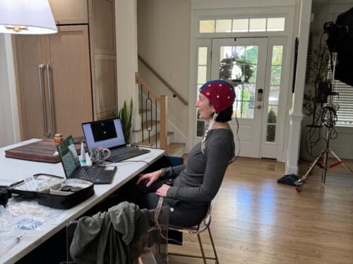 A person wearing a red cap sits sideways on a stool at a kitchen island with two laptops—one running neurofeedback software—a water bottle, and various items. Light streams in from windows, and filming equipment is set up in the background.
