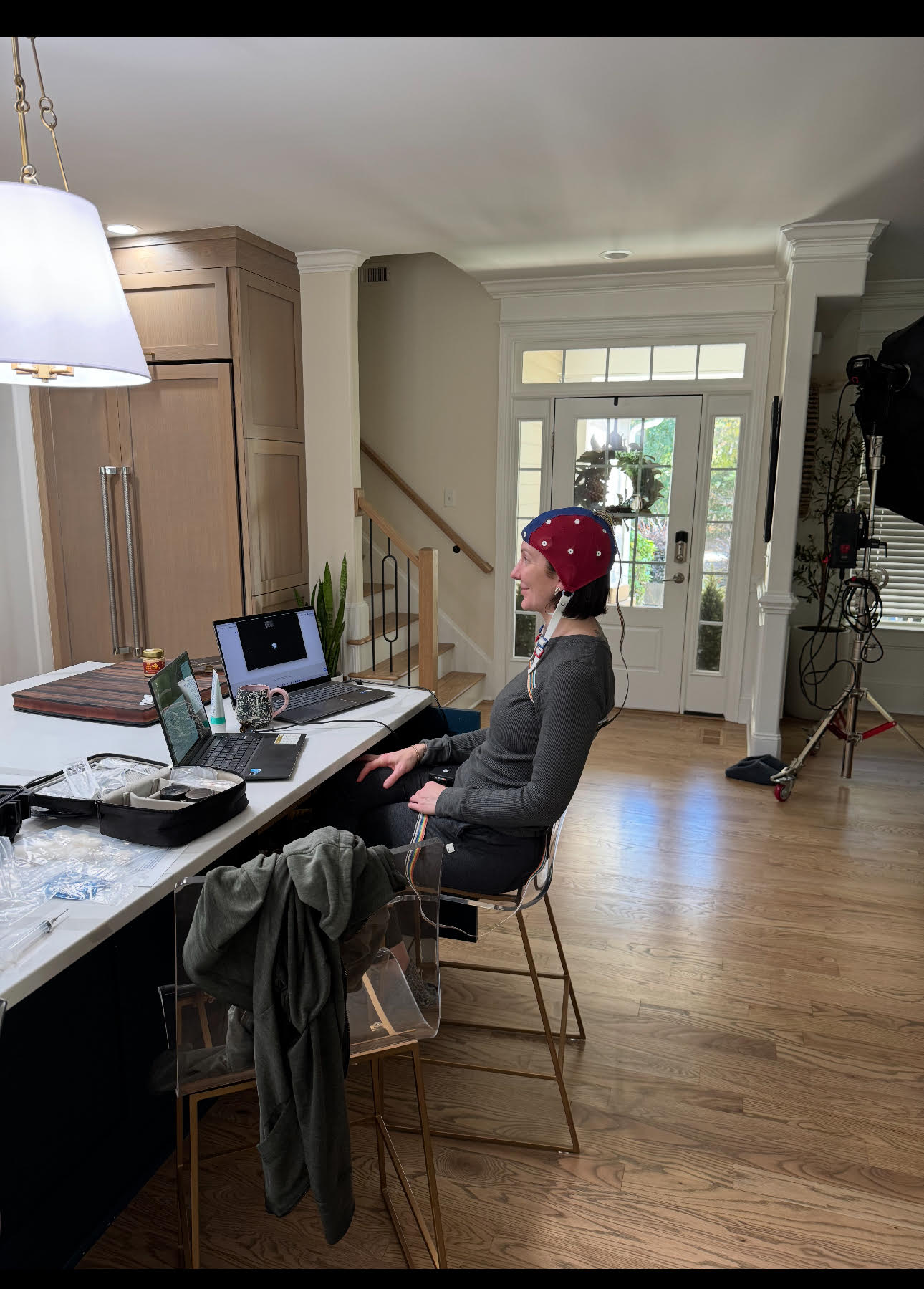 A person wearing a red cap sits sideways on a stool at a kitchen island with two laptops—one running neurofeedback software—a water bottle, and various items. Light streams in from windows, and filming equipment is set up in the background.