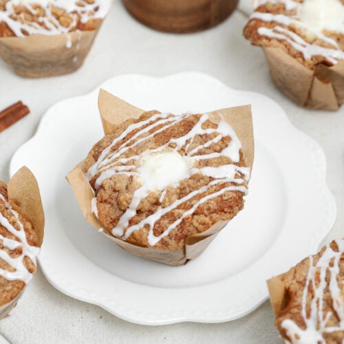 Banana bread coffee cake muffins with cinnamon streusel and vanilla glaze in parchment muffin liners on a white plate.