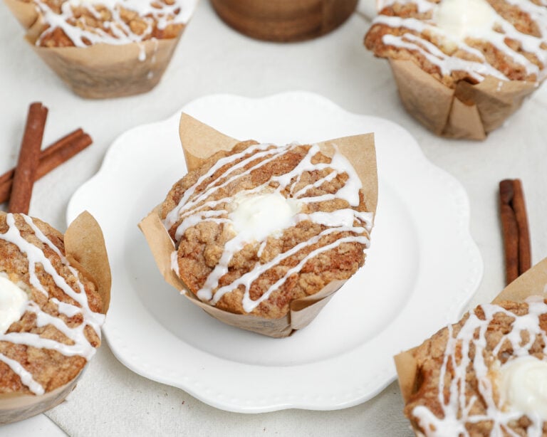 Banana bread coffee cake muffins with cinnamon streusel and vanilla glaze in parchment muffin liners on a white plate.