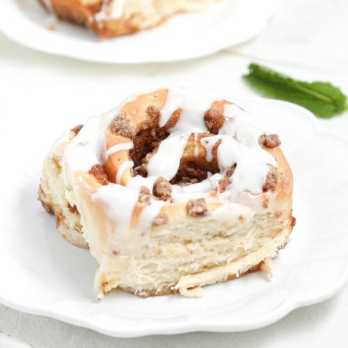Close-up of a peach cobbler cinnamon roll topped with white glaze on a white plate, showing the soft layered interior and cinnamon-peach filling.