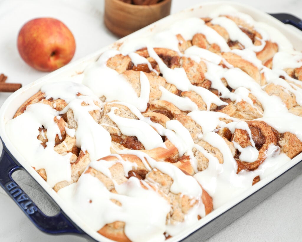 Baking dish filled with peach cobbler cinnamon rolls topped with thick white glaze, with a fresh peach and cinnamon sticks in the background.