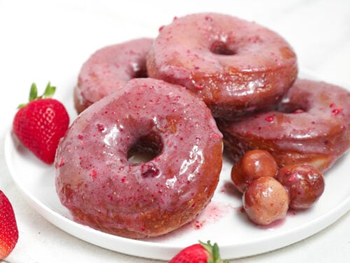 Strawberry glazed donuts stacked on a white plate with fresh strawberries and donut holes.