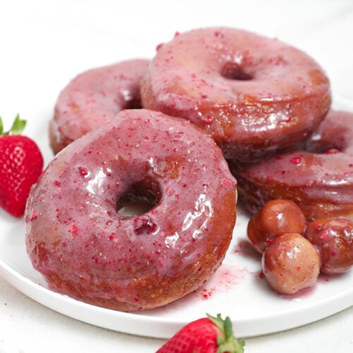 Strawberry glazed donuts stacked on a white plate with fresh strawberries and donut holes.