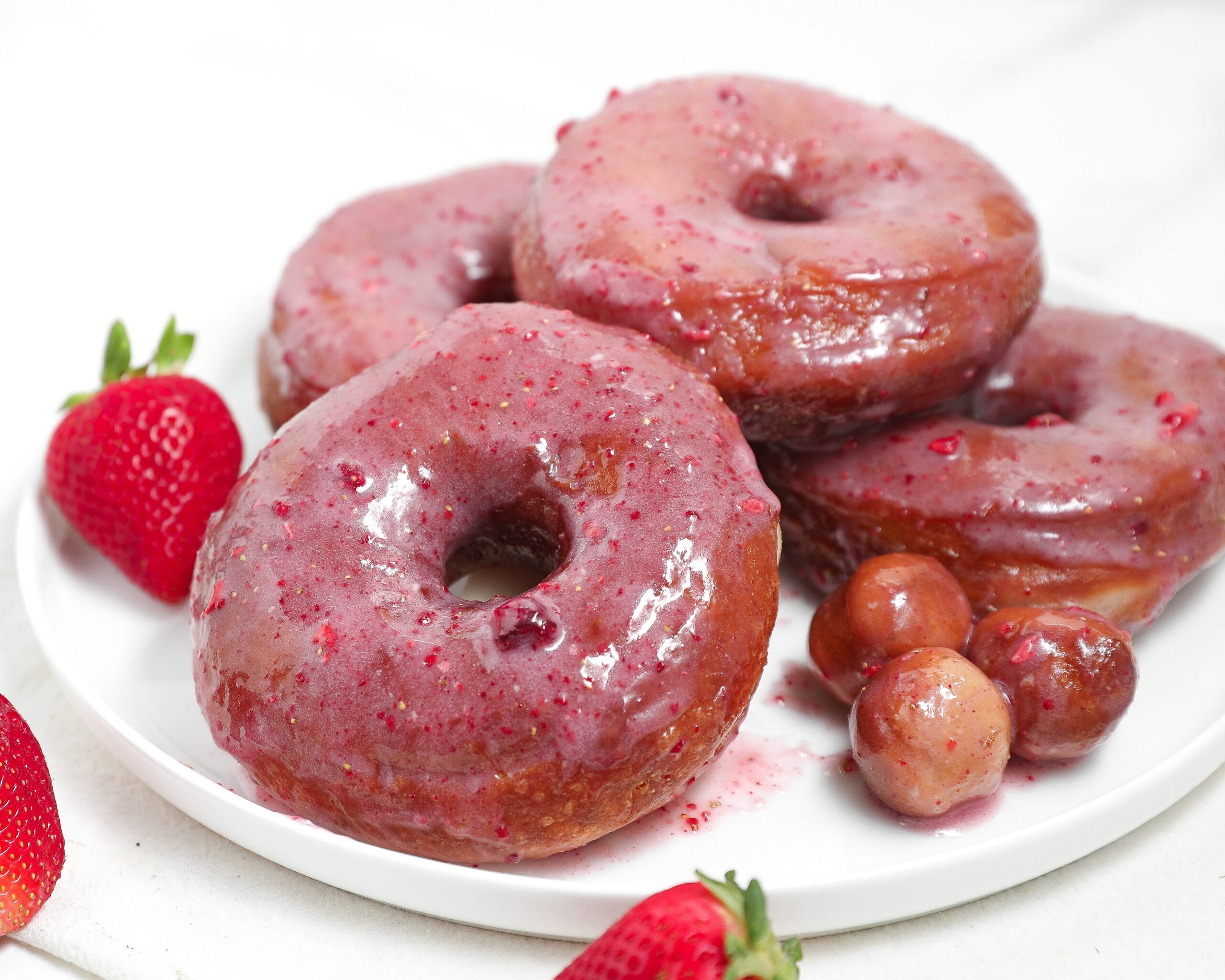 Strawberry glazed donuts stacked on a white plate with fresh strawberries and donut holes.