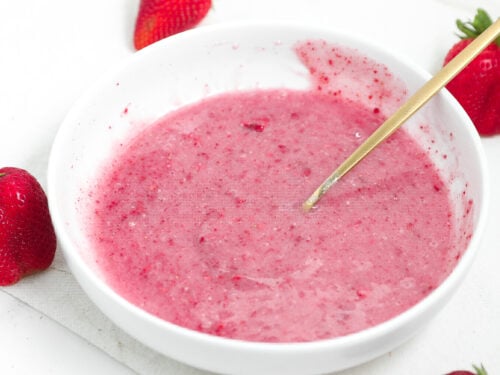 Pink strawberry glaze in a white bowl with a spoon and fresh strawberries around it.
