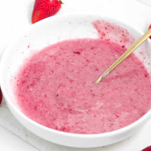 Pink strawberry glaze in a white bowl with a spoon and fresh strawberries around it.