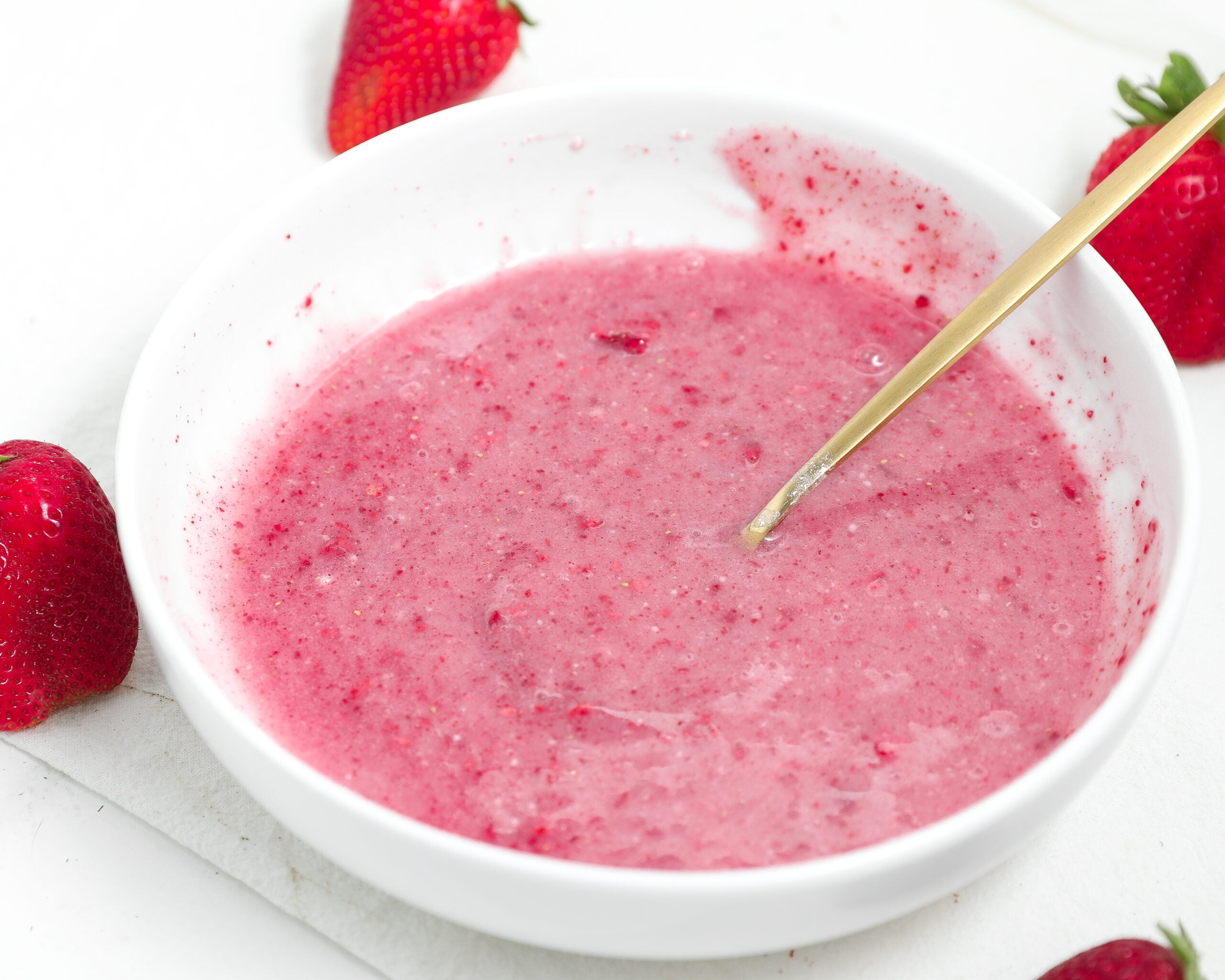 Pink strawberry glaze in a white bowl with a spoon and fresh strawberries around it.