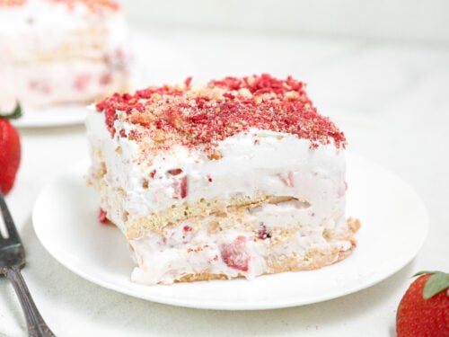 Slice of strawberry shortcake-style layered dessert with whipped cream, fresh strawberries, and cookie or cake layers, topped with strawberry crumble on a white plate