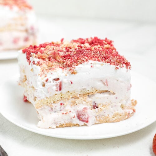 Slice of strawberry shortcake-style layered dessert with whipped cream, fresh strawberries, and cookie or cake layers, topped with strawberry crumble on a white plate