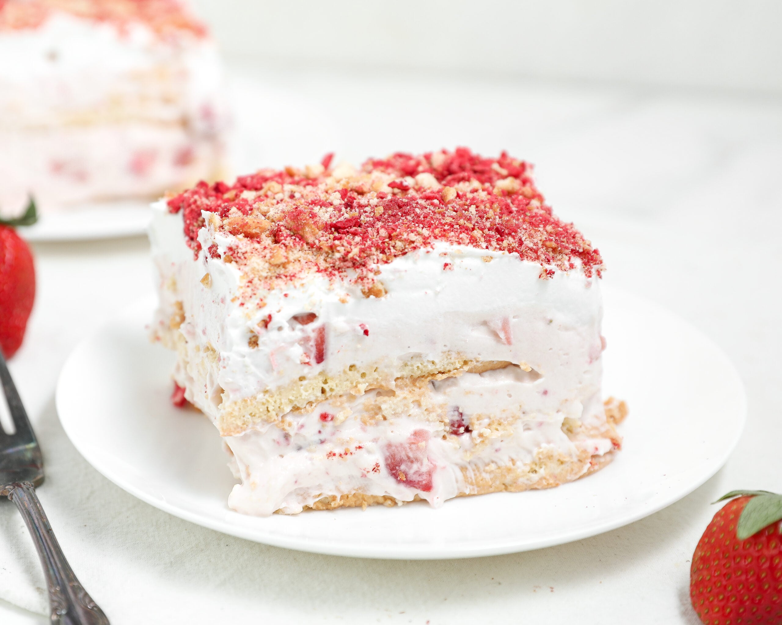 Slice of strawberry shortcake-style layered dessert with whipped cream, fresh strawberries, and cookie or cake layers, topped with strawberry crumble on a white plate