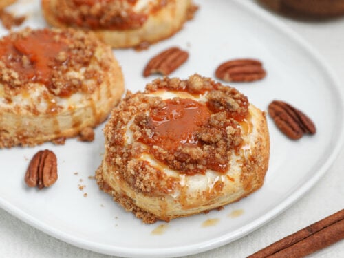 Pumpkin Cream Cheese Danish topped with Biscoff cookie crumble and caramel drizzle on a white plate with pecans and cinnamon sticks.