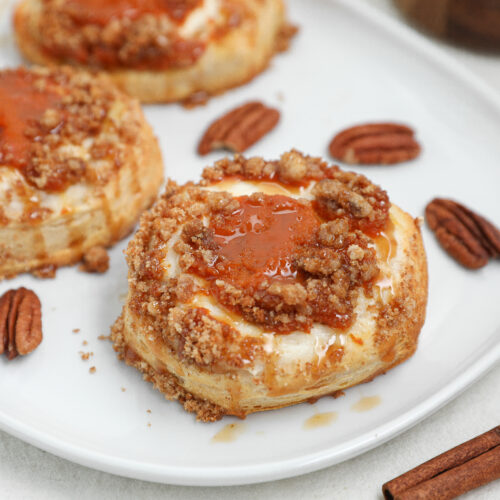 Pumpkin Cream Cheese Danish topped with Biscoff cookie crumble and caramel drizzle on a white plate with pecans and cinnamon sticks.