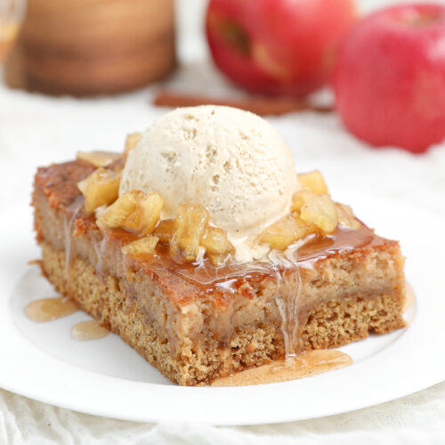 Slice of Caramel Apple Gooey Butter Cake topped with warm cinnamon caramel apples, vanilla ice cream, and caramel sauce on a white plate.