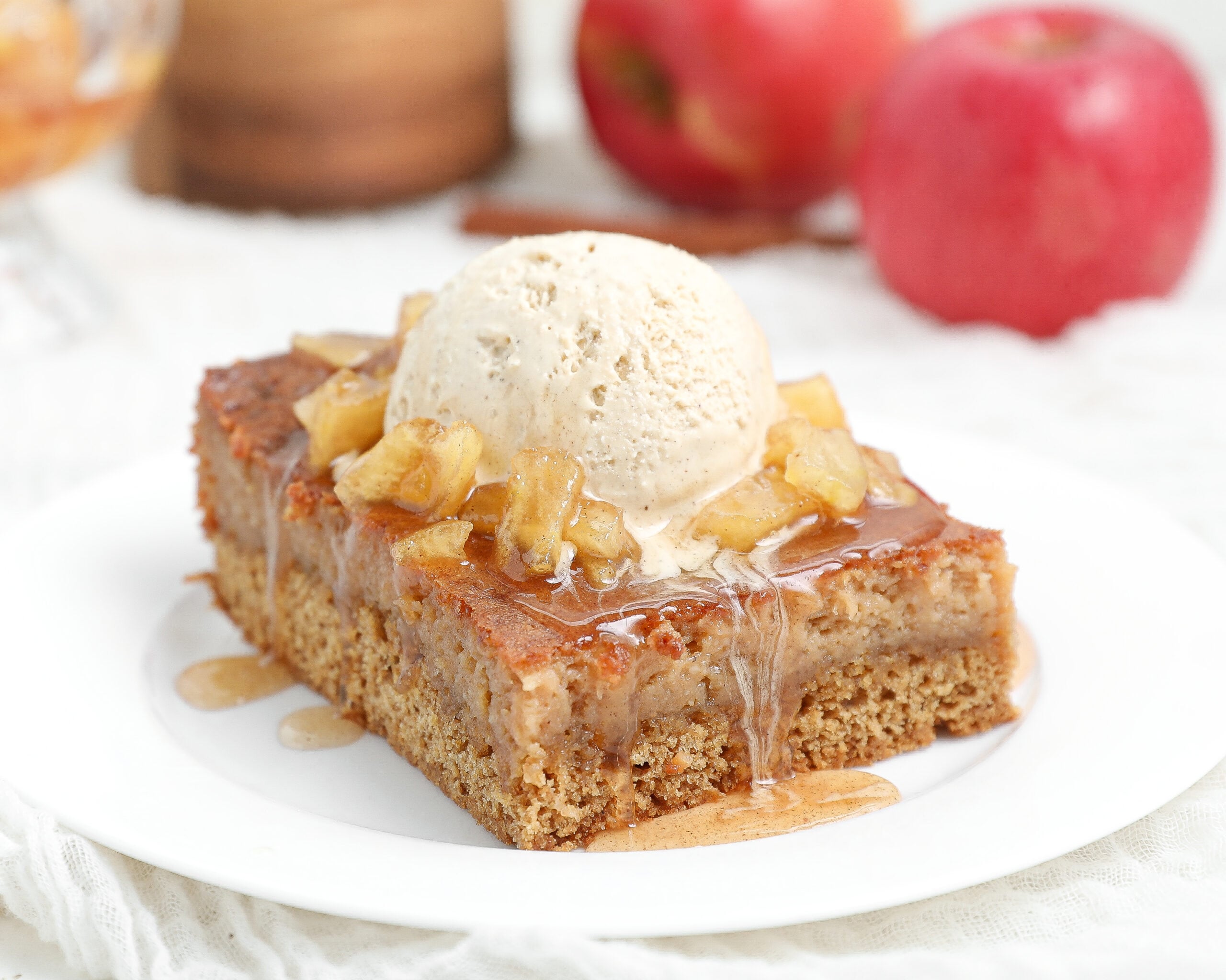 Slice of Caramel Apple Gooey Butter Cake topped with warm cinnamon caramel apples, vanilla ice cream, and caramel sauce on a white plate.