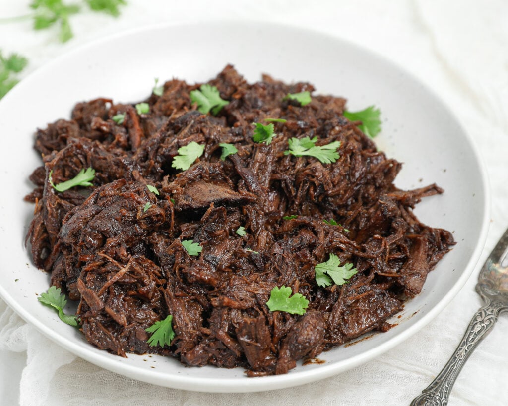 Honey Balsamic Chuck Roast shredded and served in a white bowl with fresh parsley garnish.