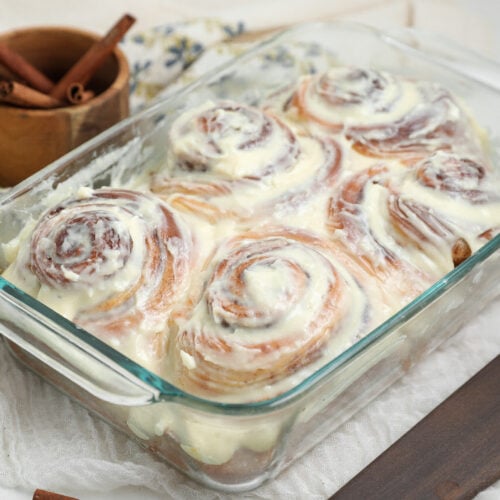 Small batch cinnamon rolls baked in a glass dish and topped with cream cheese frosting, with cinnamon sticks in the background.