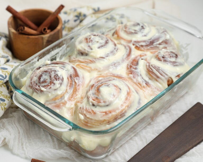 Small batch cinnamon rolls baked in a glass dish and topped with cream cheese frosting, with cinnamon sticks in the background.
