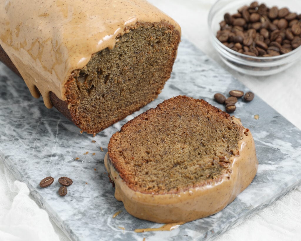 Espresso Banana Bread with a thick coffee glaze, shown as a sliced loaf on a gray marble board with coffee beans in the background.