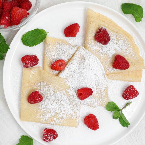Easy crepes folded into triangles on a white plate, dusted with powdered sugar and topped with fresh raspberries and mint leaves