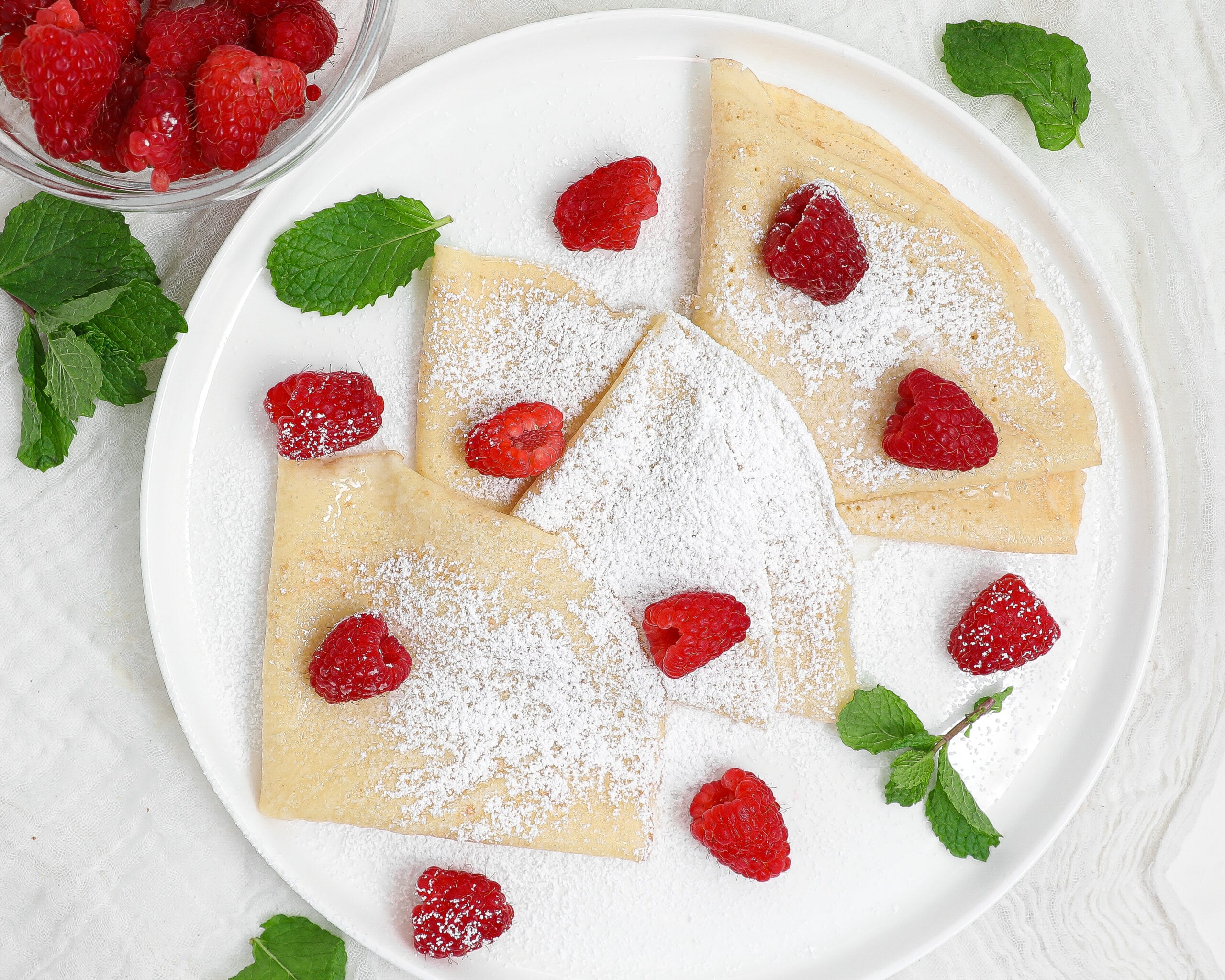 Easy crepes folded into triangles on a white plate, dusted with powdered sugar and topped with fresh raspberries and mint leaves