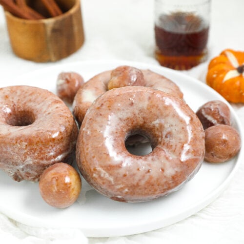 Soft pumpkin doughnuts covered in maple glaze on a white plate with pumpkin doughnut holes and fall decorations.