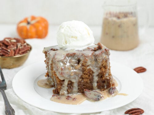 A slice of pecan praline sticky pudding cake covered in creamy pecan sauce and topped with vanilla ice cream on a white plate.
