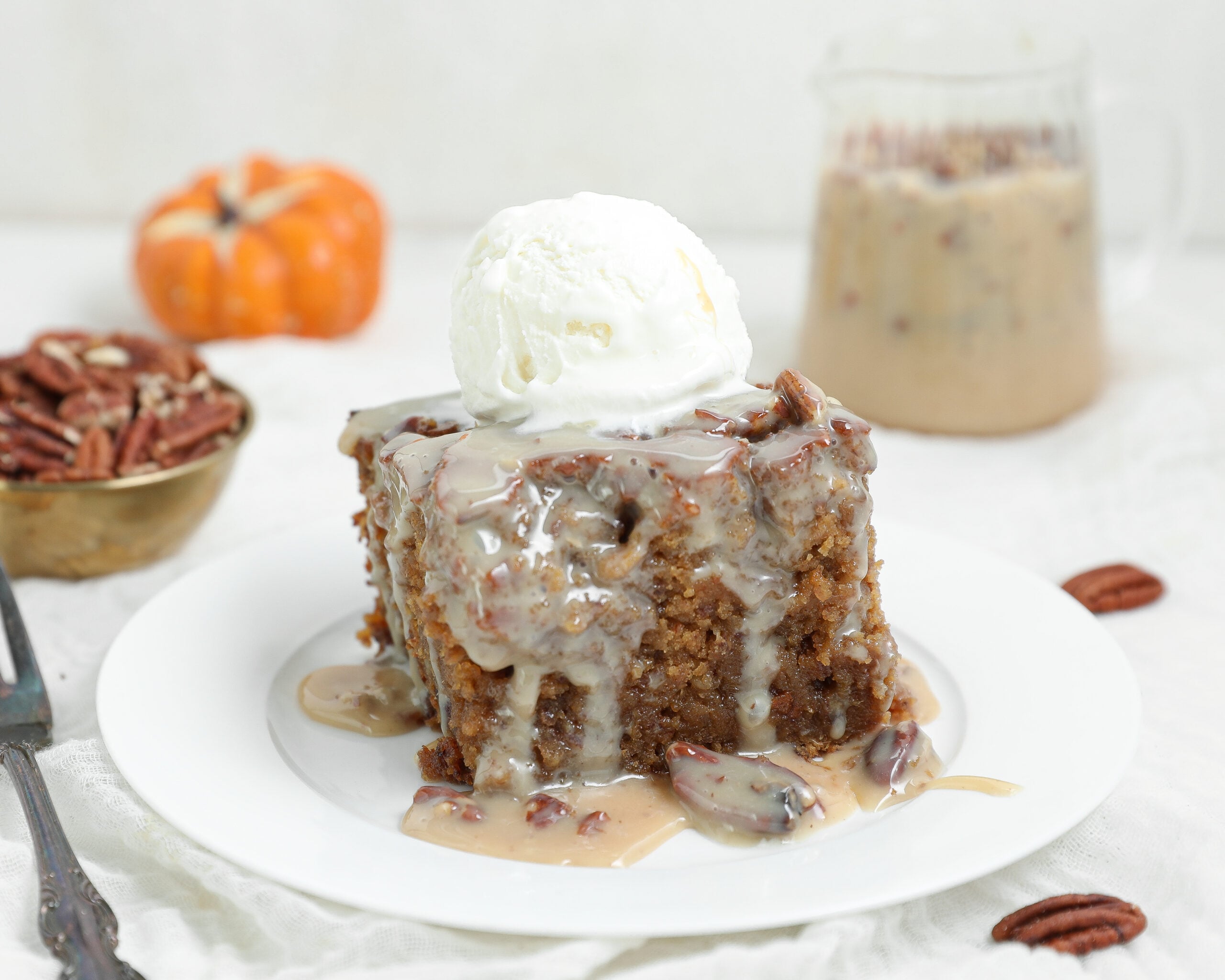 A slice of pecan praline sticky pudding cake covered in creamy pecan sauce and topped with vanilla ice cream on a white plate.