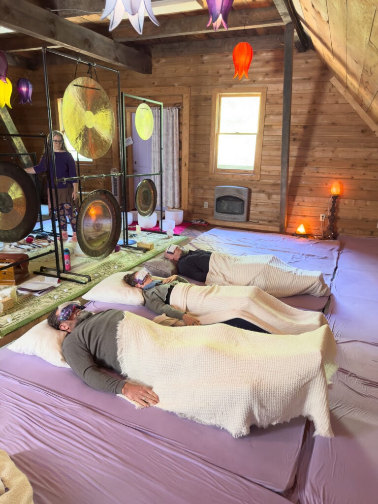 Guests resting on cushioned mats beside gongs and singing bowls in a wood-paneled sound bath room.